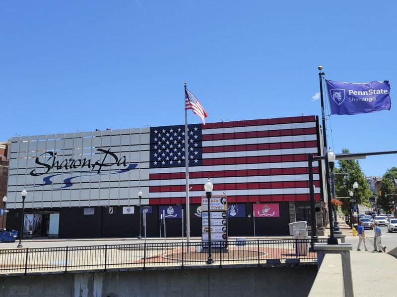 Exterior of a building with an American flag painted on the side and text saying Sharon, PA with sidewalks and flag poles in the foreground