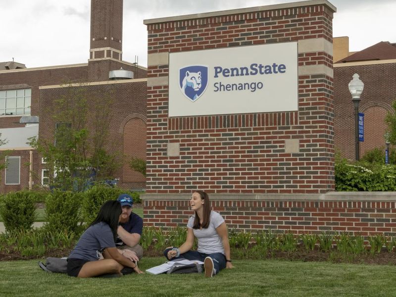 Three students sit on the ground in front of the Penn State Shenango sign