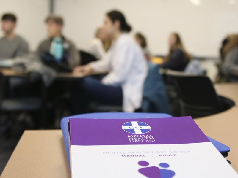 Mental Health First Aid booklet on a desk in the foreground with students out of focus in the background