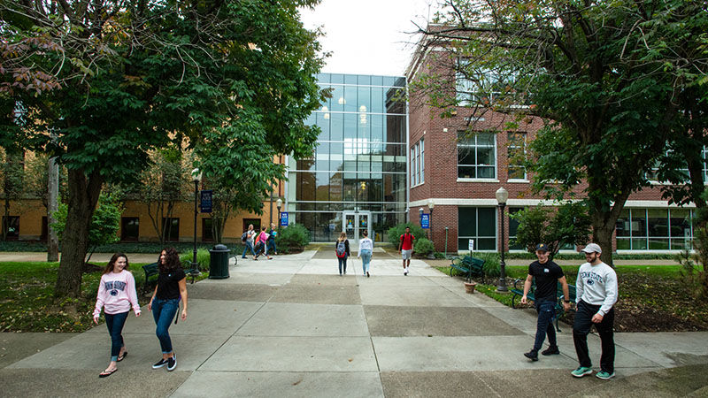Five groups of students walk on the sidewalks outside of Sharon and Lecture halls on a sunny day. 