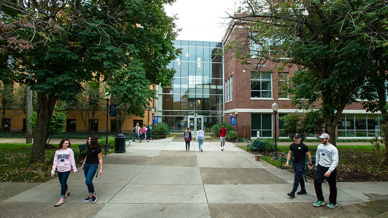 Five groups of students walk on the sidewalks outside of Sharon and Lecture halls on a sunny day.