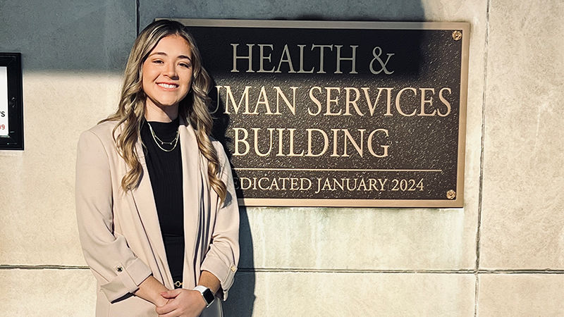 Jessica Divens posing in front of a metal sign outside of the Pennsylvania Health and Human Services building