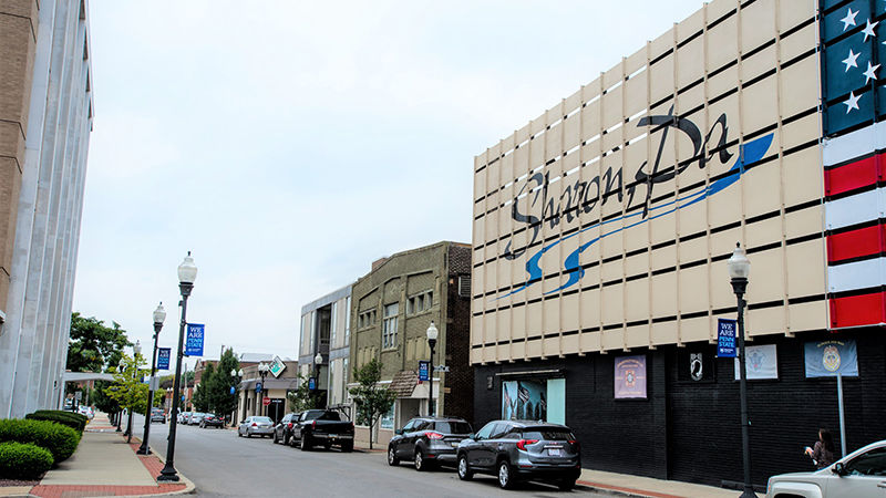 The Sharon, PA logo is displayed on two stories of a downtown building on Shenango Avenue. The street is lines with cars on one side and, on the other, lamp posts with flags that say "We are Penn State." 