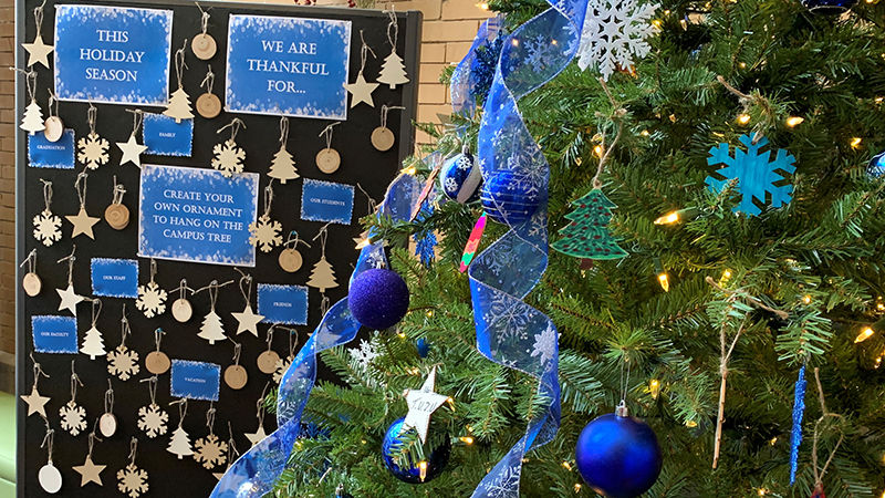 Blank ornaments in the background with signs prompting participants to write what they're thankful for. A decorated holiday tree is in the foreground. 