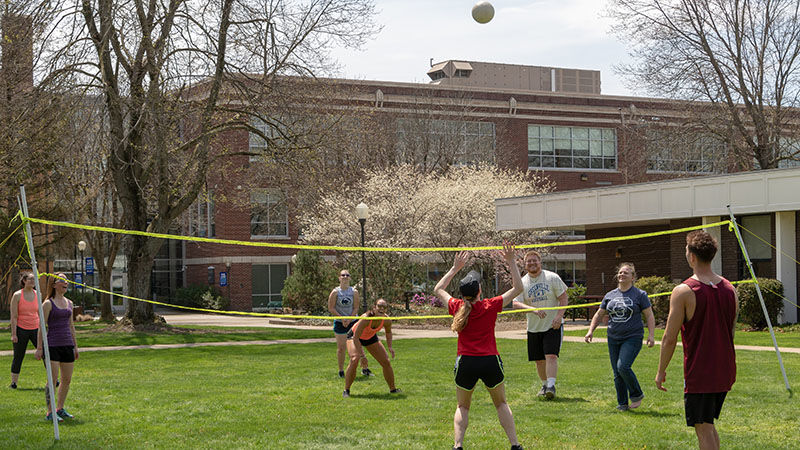 A group of eight Shenango students are hitting the volleyball high into the air and over the net on a sunny day in the grass in front of Forker Lab on campus. 