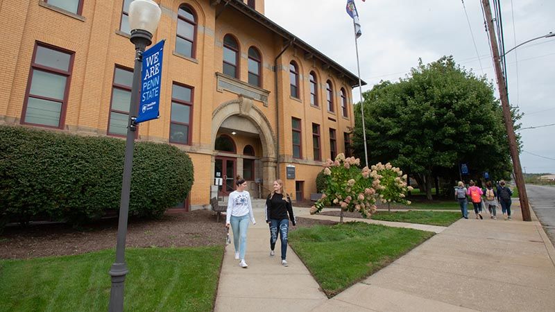 Two students walk out of Lecture Hall on a sidewalk while four other students walk along Shenango Avenue. 