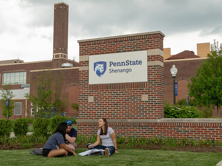 3 students sitting in grass in front of campus sign