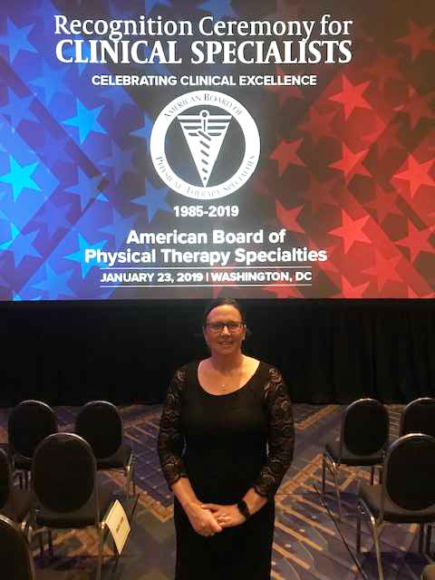 A woman standing in front of a projected image that reads recognition ceremony for clinical specialists celebrating clinical excellence 1985 to 2019 American Board of Physical Therapy Specialities January 23 2019 Washington DC