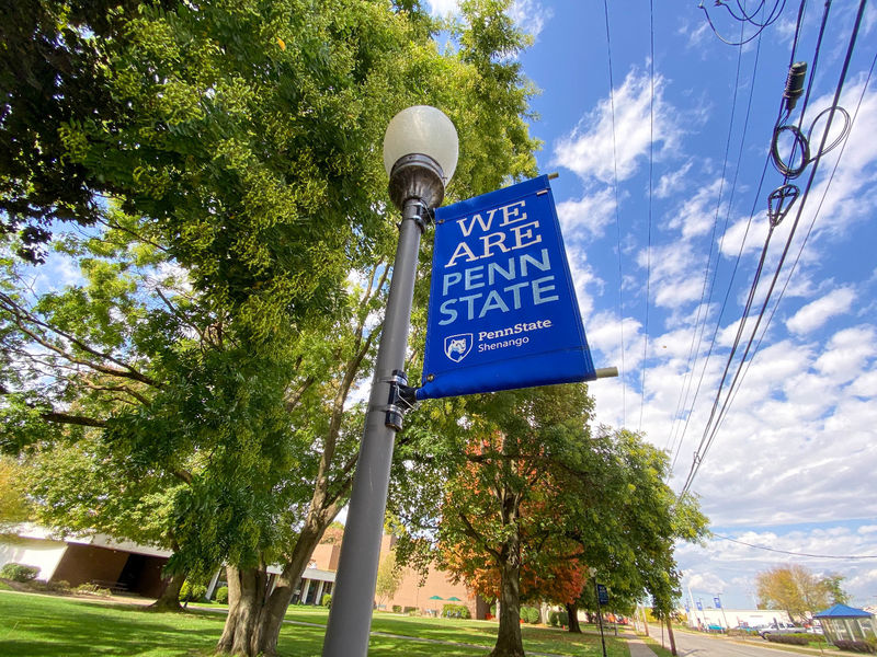 "We are Penn State" sign on lamp pole with green and orange trees in background