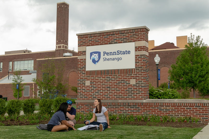 Three students study in front of the front entrance sign, which features Penn State logo, with the Auditorium in the background. 
