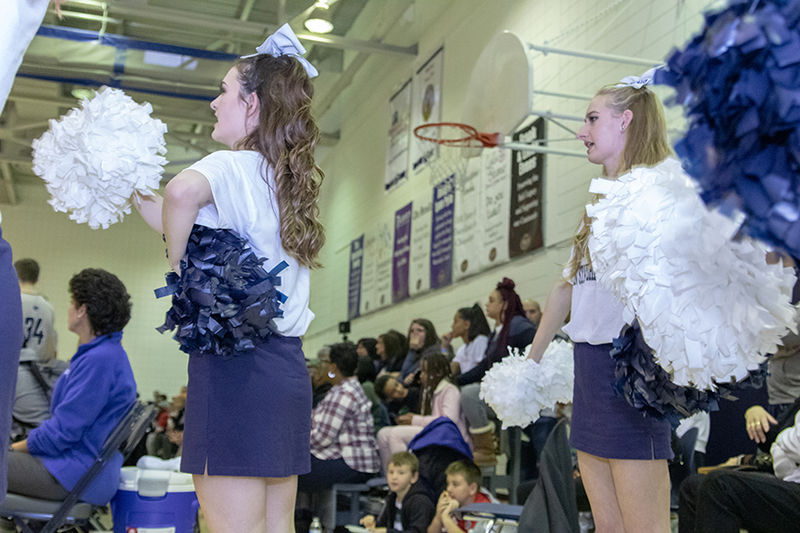 Two cheerleaders stand to the side of the bleachers and waive a pom-pom in each hand. 