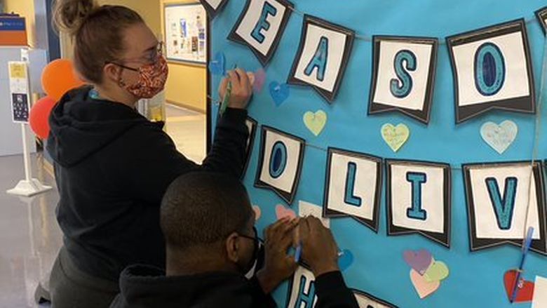 Two students writing on hearts pinned to a board titled "100 reasons to live"