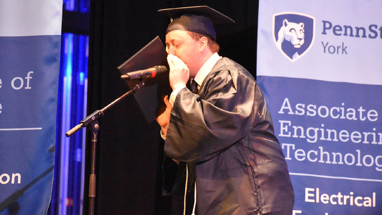 Male student in graduation cap and gown speaking into a microphone on a stabnd