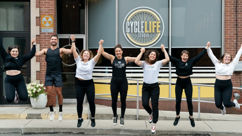 The CycleLife Studio team holds hands and jumps in unison on the street outside of the CycleLife Studio location