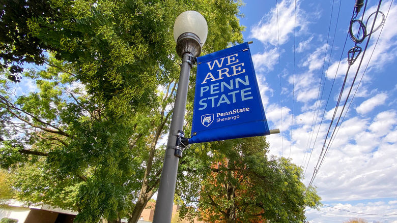 "We are Penn State" sign on lamp pole with green and orange trees in background