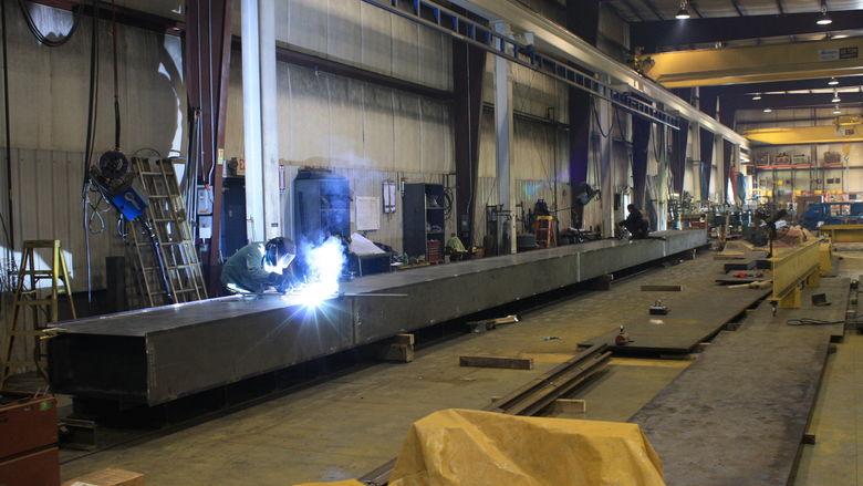 Factory employees work on a metal beam. One employee is welding the metal. 