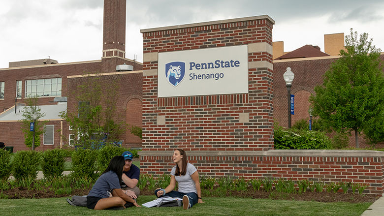 Three students study in the grass at the entrance to campus, in front of the "Penn State Shenango" welcome sign. 