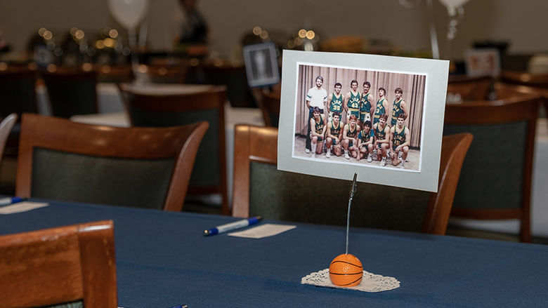 On a tablescape, a printed group photo of an alumni team of men's basketball players sits on a blue tablecloth with place settings. 