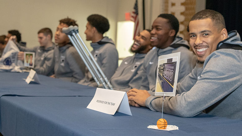 The men's basketball team sits at a long table at the athletic alumni event. The men laugh and smile as they prepare. 