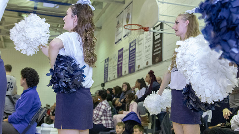 Two cheerleaders stand to the side of the bleachers and waive a pom-pom in each hand. 