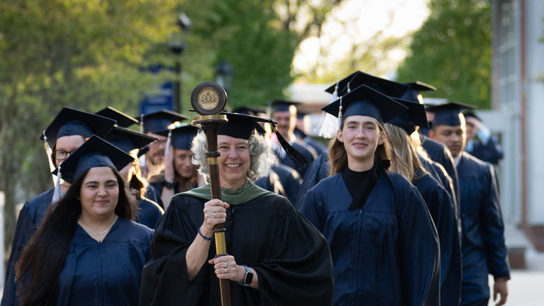 Graduates in caps and gowns in two rows walking down sidewalk led by faculty member in black robe carrying a staff and pendant.