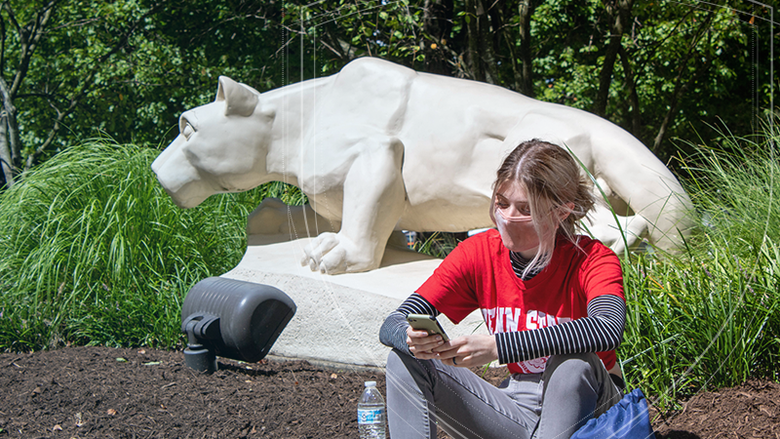Female student in red shirt and mask sits on steps in front of the Nittany Lion Shrine