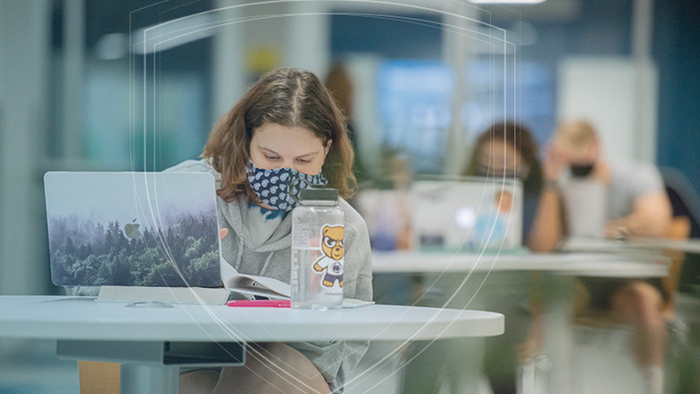 A female student works on a laptop in a student lounge. She and other students in the background are wearing masks and social distancing. 