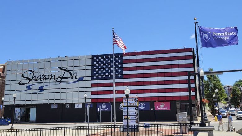 Exterior of a building with an American flag painted on the side and text saying Sharon, PA with sidewalks and flag poles in the foreground