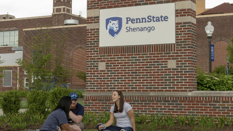Three students sit on the ground in front of the Penn State Shenango sign