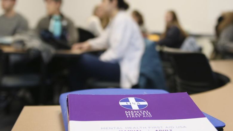 Mental Health First Aid booklet on a desk in the foreground with students out of focus in the background