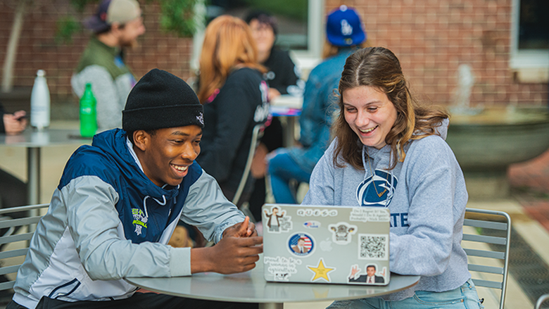 Two students working on laptop in a campus courtyard. 