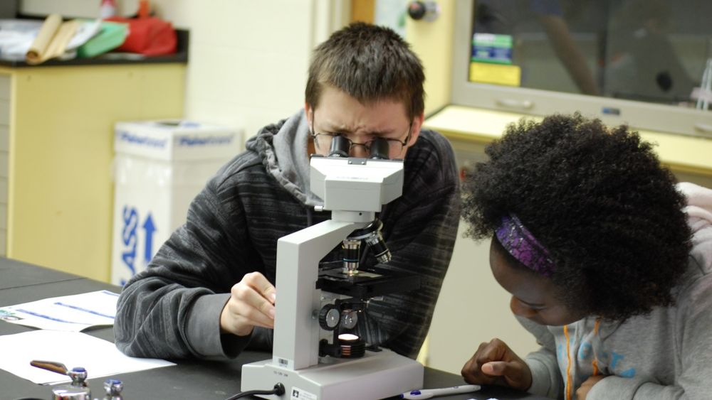 Two young students are pictured--one looking into a microscope and the other taking notes.