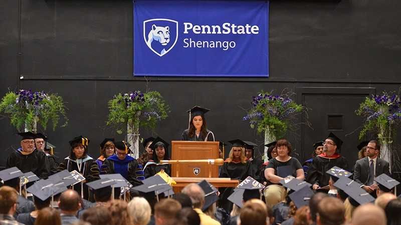A student stands at a podium speaking while other faculty and staff sit on the stage watching.