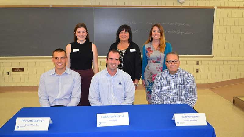 Three men are sitting at a table while three women are standing behind them.