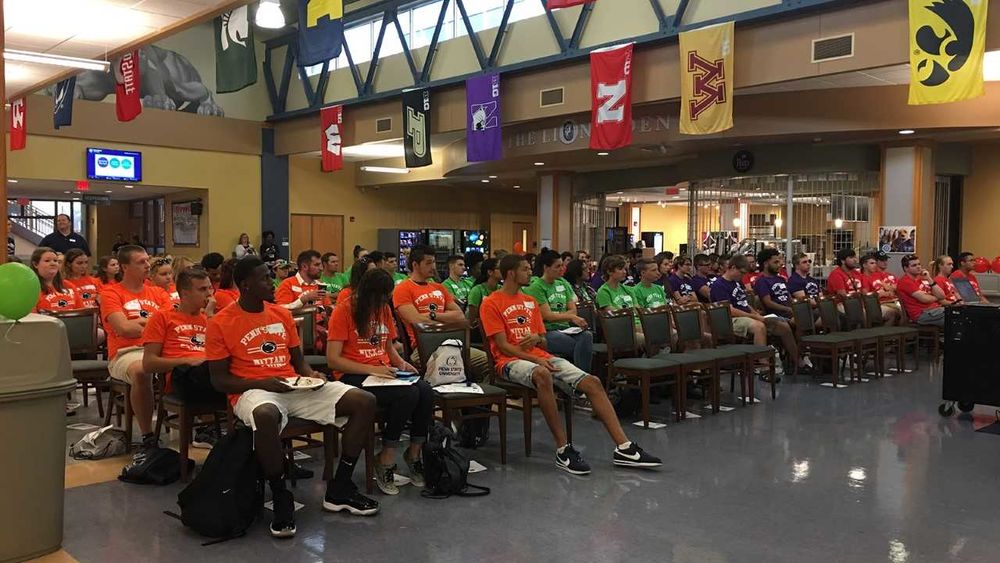 Approximately one hundred new students are seated in the campus's Great Hall wearing various colors of T-shirts.