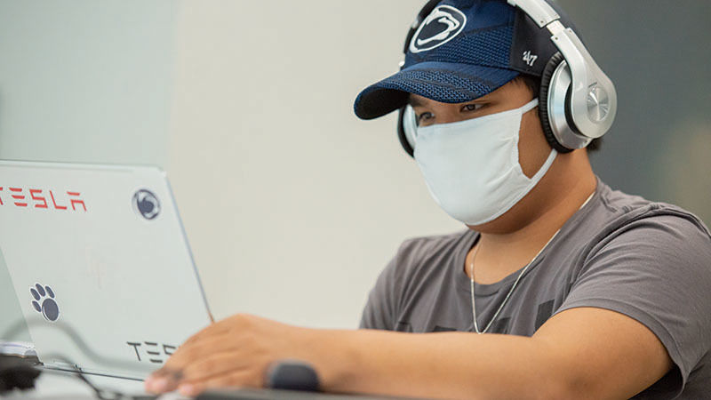 Male student in Penn State hat and headphones works on laptop in a student lounge. 