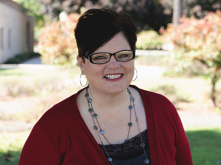 Headshot of Professor Carol Cowan-Crawford wearing a red shirt and smiling at the camera in front of a blurred out background of trees and bushes