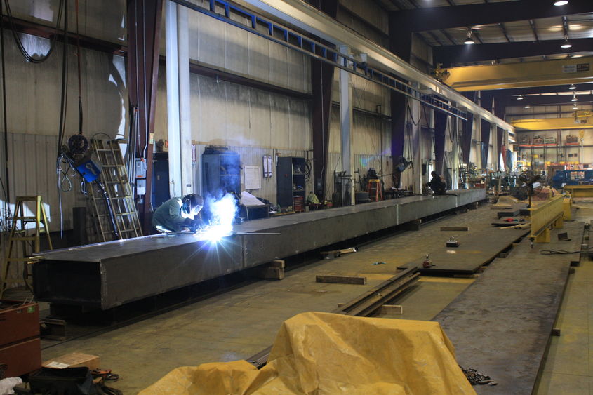 Factory employees work on a metal beam. One employee is welding the metal. 