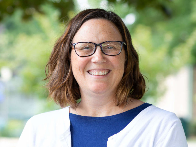 Headshot of Professor Jennifer Jewell wearing a blue shirt with a white sweater standing in front of trees