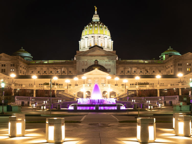Exterior view of the Pennsylvania State Capitol Building at night with lights and a fountain illuminated in a purple light