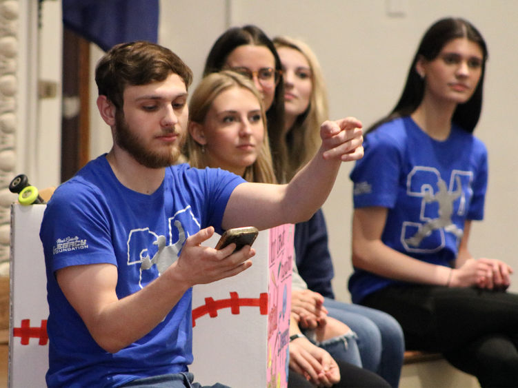 Students sitting on a stage with one looking at a phone and pointing to a crowd off camera