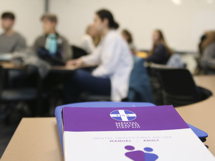 Mental Health First Aid booklet on a desk in the foreground with students out of focus in the background