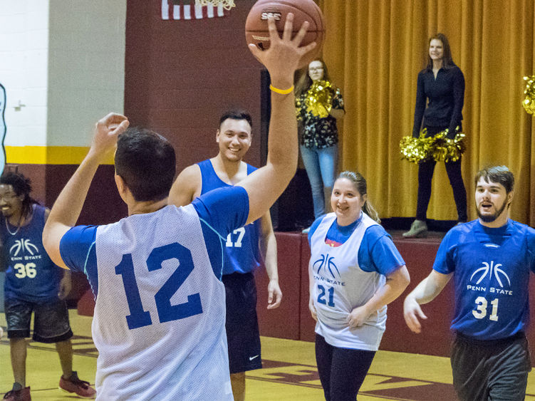 Five people wearing jerseys on a basketball court. One is facing away from the camera and holding the ball in the air while the others look and smile