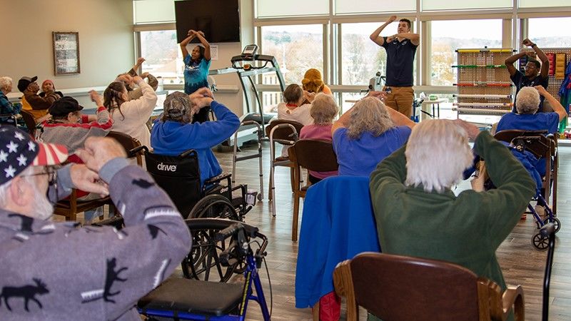 A group of seniors complete exercises in their chairs while instructors stand in front of room